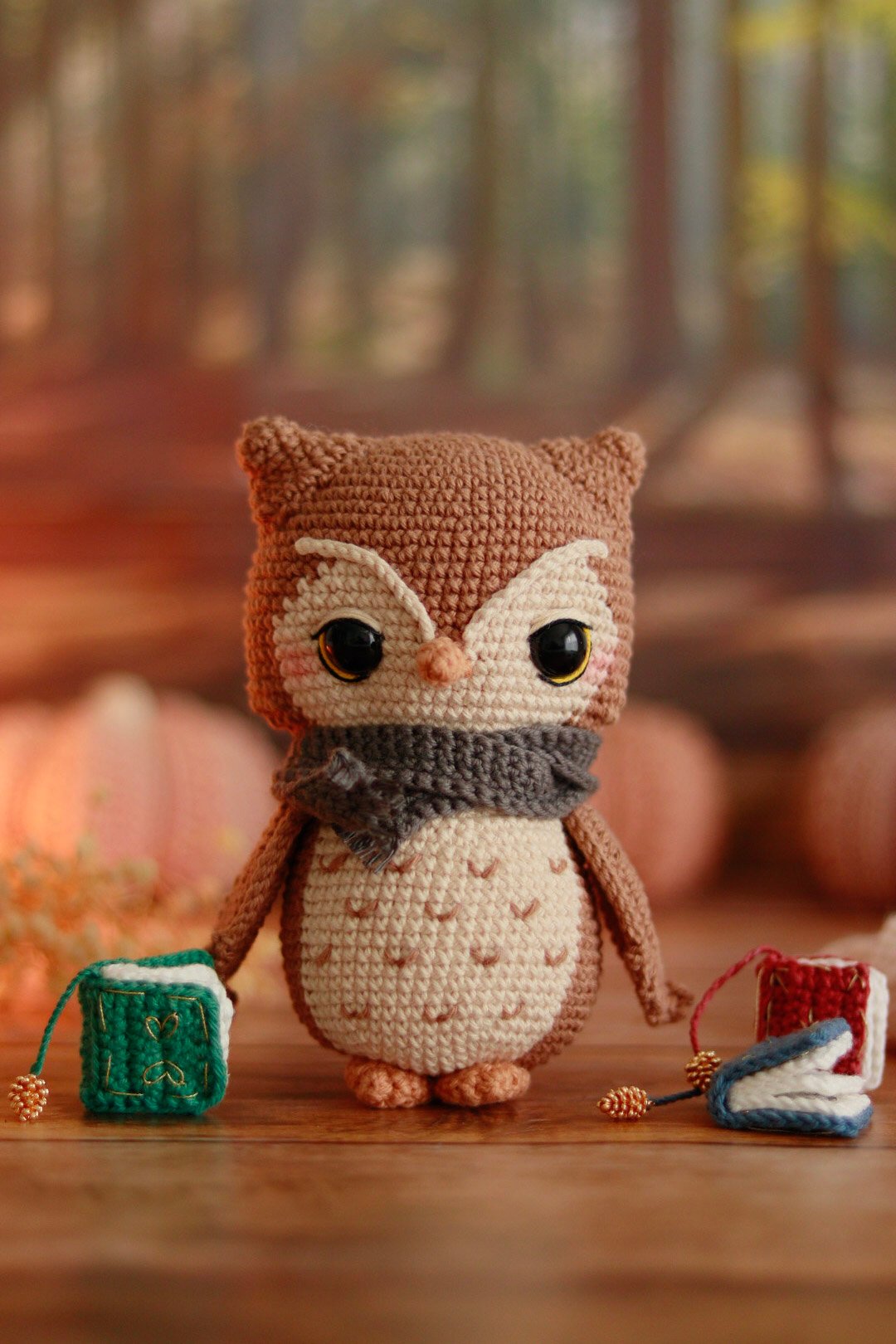 Amigurumi hibou marron et crème avec écharpe grise, tenant un mini grimoire vert au crochet dans un décor d'automne